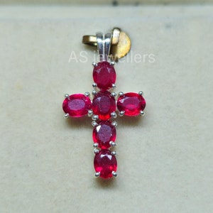 Puede incluir: Un colgante de cruz de plata con siete piedras preciosas ovaladas rojas. El colgante está sobre un fondo blanco.