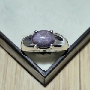May include: A silver ring featuring a large, oval, star-shaped gemstone in shades of purple and gray. The gemstone is held in place by four small prongs. The ring has a smooth, polished band.