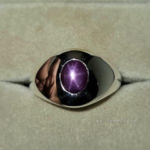 May include: A silver ring with a large, oval, purplish-red star ruby gemstone. The ring has a smooth, rounded band and a polished finish. The text "as jewellers" is visible at the bottom of the image.