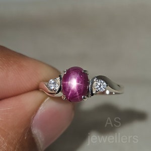 Peut inclure: Une bague en argent avec une pierre précieuse en rubis étoilé rose et deux petites pierres blanches. La bague est sertie sur une bande simple.