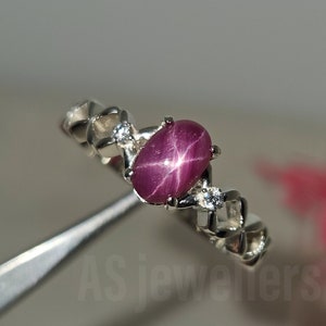 Peut inclure: Une bague en argent avec une pierre précieuse en rubis étoilé rouge et de petits diamants blancs. La bague est sertie dans un design délicat et complexe.