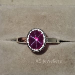 Pode incluir: Um anel de prata com uma gema de rubi estrela rosa. O rubi tem um padrão em forma de estrela no centro. O anel está em uma pequena caixa com o texto "AS jewellers" na parte inferior.