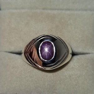 Pode incluir: Um anel de prata com uma gema de safira estrelada roxa. O anel tem um design detalhado com uma borda elevada em torno da pedra.