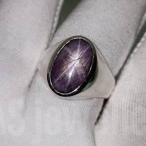 Puede incluir: Un anillo de plata con una piedra preciosa de zafiro estrellado púrpura. La piedra preciosa tiene un patrón en forma de estrella.