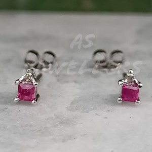 May include: A pair of silver stud earrings with a small square-shaped pink gemstone in the center of each earring.
