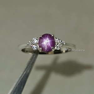 May include: A silver ring with a central, oval, star-shaped, purple gemstone. The ring is accented with smaller, clear gemstones. The ring is displayed against a neutral backdrop.