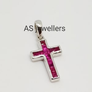 Puede incluir: Un colgante de cruz de plata con una incrustación de piedra de rubí rojo. AS Jewellers está impreso en el fondo blanco.
