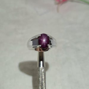 May include: A silver ring featuring a polished, oval-shaped deep red star ruby gemstone set in a four-prong mounting. The band is smooth and metallic, showcasing a distinct six-rayed star effect within the center of the dark red cabochon stone.
