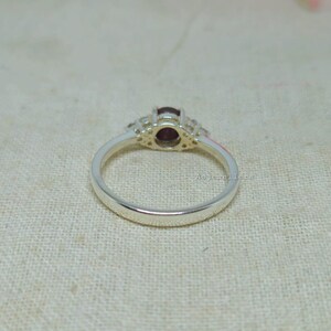 May include: A silver ring with a central oval, dark red gemstone, flanked by smaller, clear stones. The ring band is smooth and polished. The ring is set against a neutral, textured background.