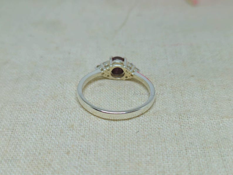 May include: A silver ring with a central oval, dark red gemstone, flanked by smaller, clear stones. The ring band is smooth and polished. The ring is set against a neutral, textured background.