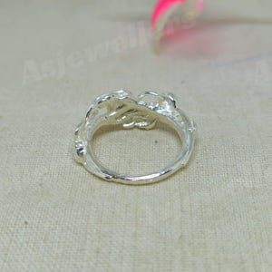 May include: A silver ring with an ornate, floral design. The ring features a detailed, textured pattern around the band and a cluster of stylized flowers at the top. The ring is shown on a neutral, textured background.