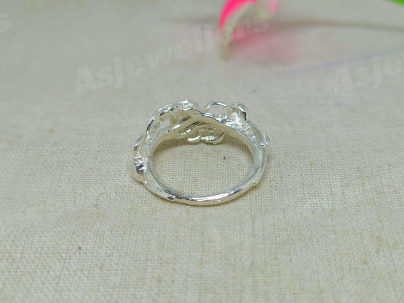 May include: A silver ring with an ornate, floral design. The ring features a detailed, textured pattern around the band and a cluster of stylized flowers at the top. The ring is shown on a neutral, textured background.