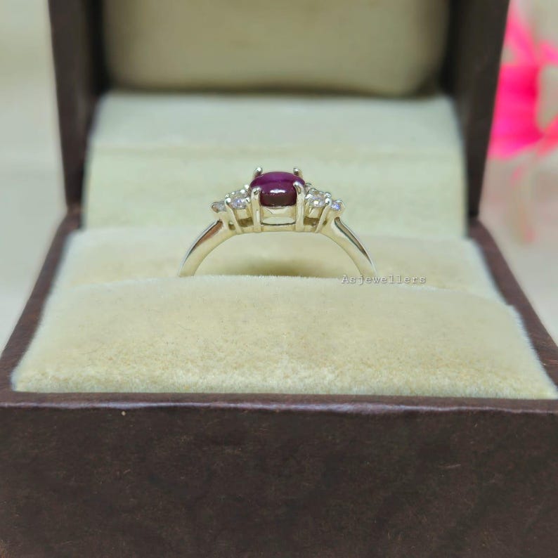 May include: A silver ring with a large oval ruby gemstone as the centerpiece, flanked by smaller, clear gemstones. The ring is presented in a brown velvet-lined jewelry box, creating a luxurious display.