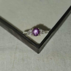 May include: A silver ring featuring a large, oval, star-shaped purple gemstone as the centerpiece. The ring is flanked by smaller, clear gemstones on each side, set in a delicate band. The ring is displayed in a dark box.