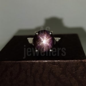 May include: A silver ring featuring a large, oval-shaped, purple star ruby gemstone. The gemstone has a star-like pattern in the centre.