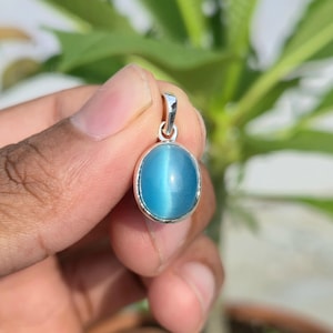 Puede incluir: Un colgante de forma ovalada con una piedra ojo de gato de color azul claro lisa, engastada en un marco de metal plateado pulido. El colgante incluye una pequeña anilla de metal plateado integrada en la parte superior para sujeción.
