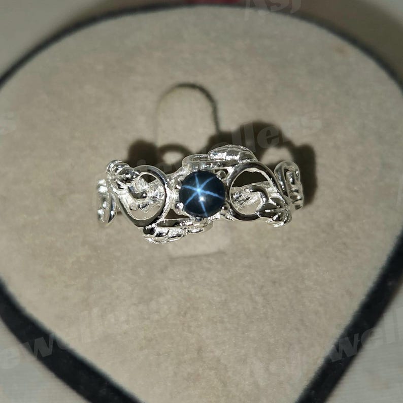 May include: A silver ring with an elaborate, swirling pattern. The ring has a dark blue, star-shaped gemstone in the middle. The ring is shown on a grey surface.
