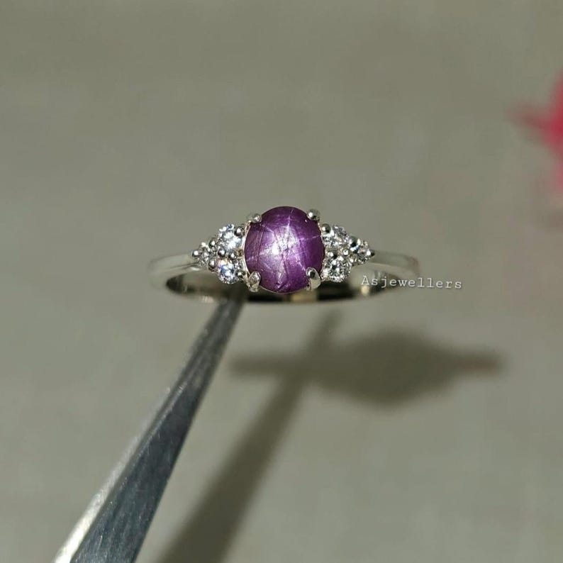 May include: A silver ring featuring a large, oval, star-shaped, purple gemstone. The ring is flanked by smaller, clear gemstones on each side. The ring is set against a neutral background.