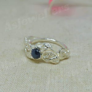 May include: A silver ring featuring a dark blue gemstone. The ring has an intricate, textured design with floral or organic shapes. The gemstone is round and centrally set.