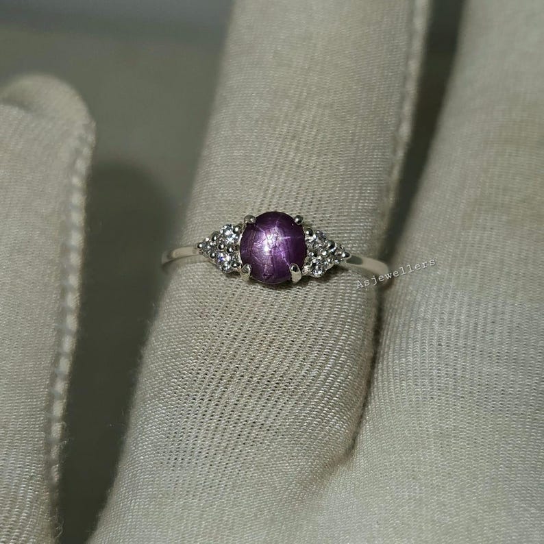 May include: A silver ring featuring a large, oval, purplish-red gemstone at the center, flanked by smaller, clear gemstones. The ring is set on a light-colored fabric. The text "Asjewellers" is visible on the ring.
