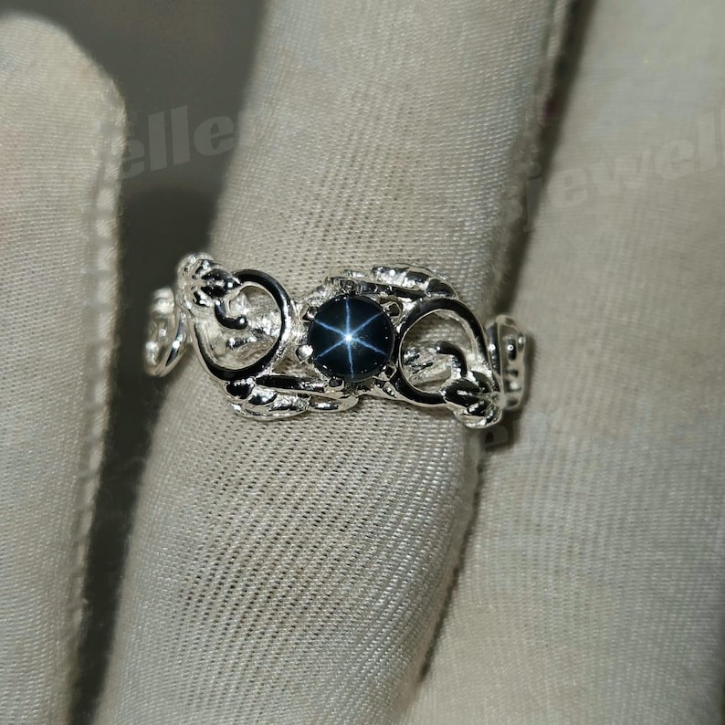 May include: A silver ring with a dark blue star sapphire. The ring has an intricate, swirling pattern around the gemstone. The ring is shown against a light grey fabric background.