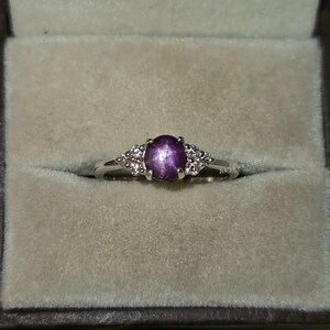 May include: A silver ring with a large oval, purple gemstone at the center, flanked by smaller, clear stones. The ring is set in a light-colored box, showcasing the jewelry's design and color.