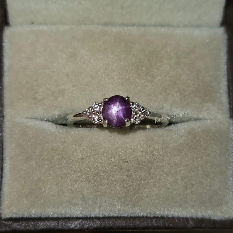 May include: A silver ring with a large oval, purple gemstone at the center, flanked by smaller, clear stones. The ring is set in a light-colored box, showcasing the jewelry's design and color.