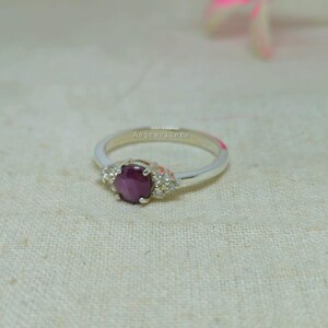 May include: A silver ring with a large, oval, deep red gemstone at the center, flanked by smaller, clear stones. The ring band is thin and polished, with the text "Asjewellers" inscribed on the band.