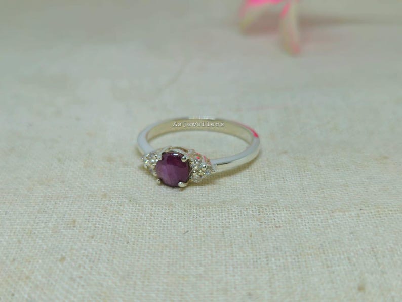 May include: A silver ring with a large, oval, deep red gemstone at the center, flanked by smaller, clear stones. The ring band is thin and polished, with the text "Asjewellers" inscribed on the band.