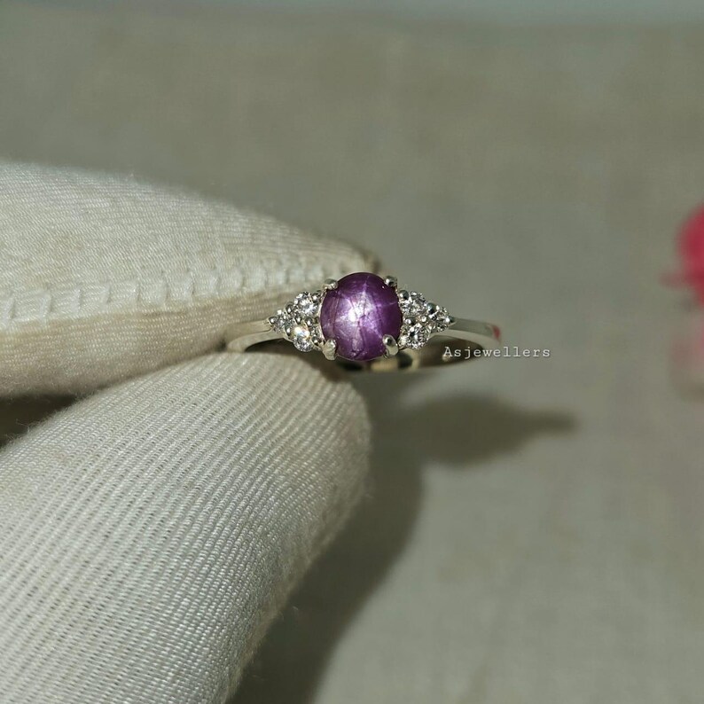 May include: A silver ring featuring a large, oval, purplish-pink gemstone with a star-like effect. The ring is flanked by smaller, clear gemstones on each side. The text "Asjewellers" is visible on the right side of the ring.