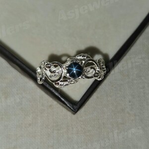 May include: A silver ring with an intricate, swirling pattern. The ring has a dark blue, star-shaped gemstone set in the centre. The ring is displayed on a dark surface.