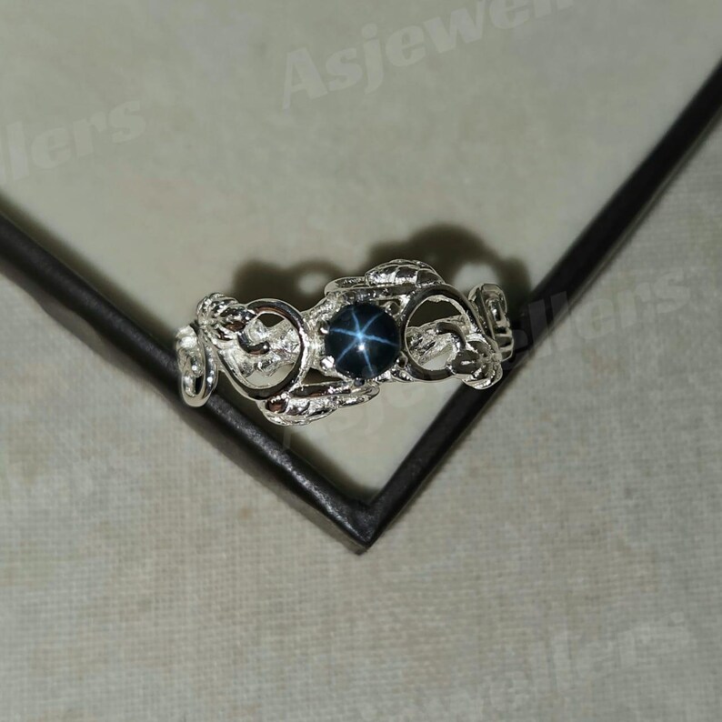 May include: A silver ring with an intricate, swirling pattern. The ring has a dark blue, star-shaped gemstone set in the centre. The ring is displayed on a dark surface.