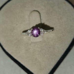 May include: A silver ring featuring a central oval, star-shaped, purple gemstone, flanked by clusters of smaller, clear gemstones. The ring is set against a gray velvet surface.