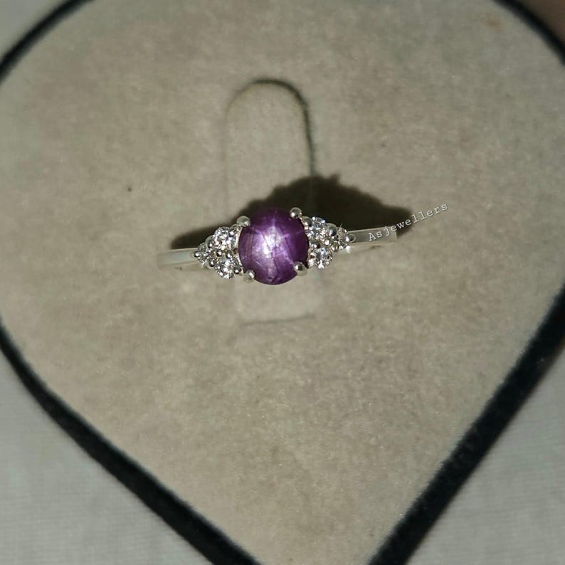 May include: A silver ring featuring a central oval, star-shaped, purple gemstone, flanked by clusters of smaller, clear gemstones. The ring is set against a gray velvet surface.