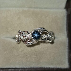 May include: A silver ring with an elaborate, openwork design. The ring features a dark blue star sapphire gemstone at its centre. The band has a flowing, organic pattern, creating a unique and elegant piece of jewellery.