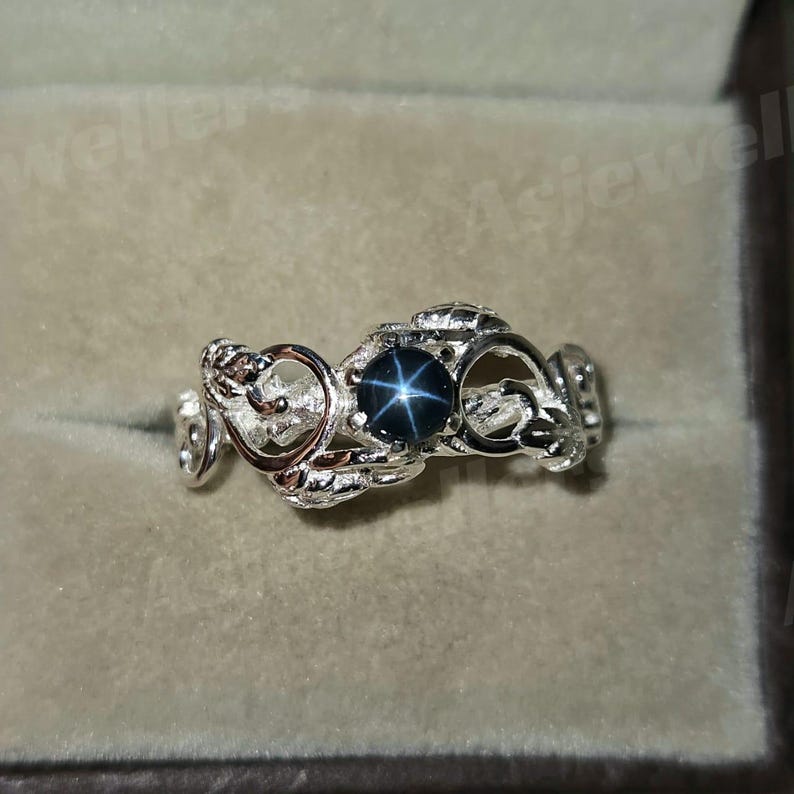 May include: A silver ring with an elaborate, openwork design. The ring features a dark blue star sapphire gemstone at its centre. The band has a flowing, organic pattern, creating a unique and elegant piece of jewellery.