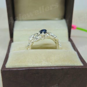 May include: A silver ring featuring a dark blue gemstone, set within an elaborate floral design. The ring is presented in a brown jewellery box with a beige interior, highlighting the ring's intricate details and craftsmanship.
