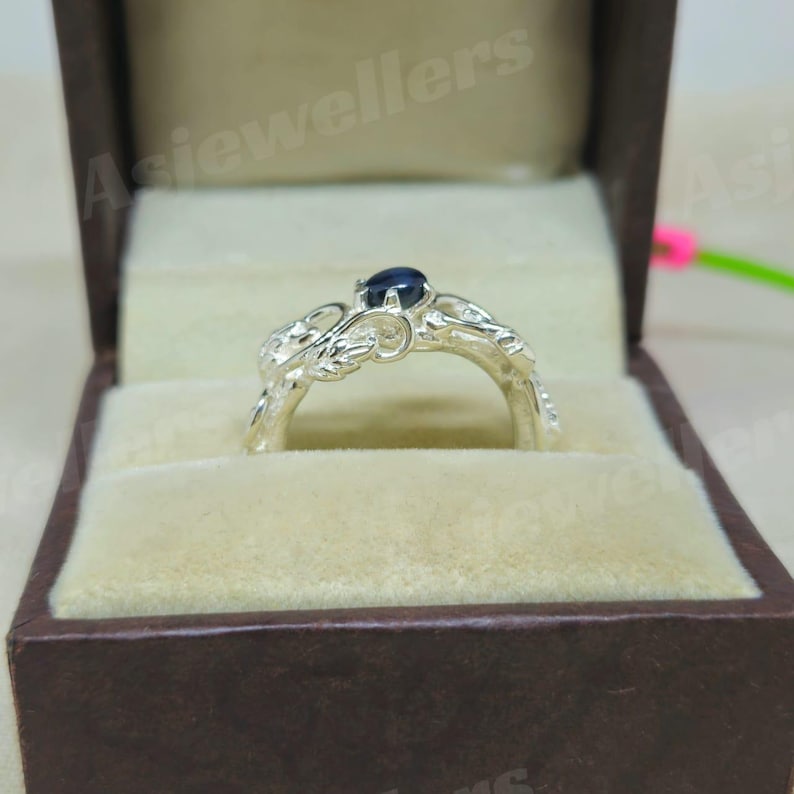 May include: A silver ring featuring a dark blue gemstone, set within an elaborate floral design. The ring is presented in a brown jewellery box with a beige interior, highlighting the ring's intricate details and craftsmanship.