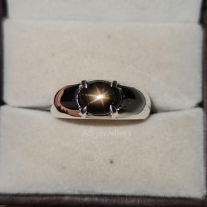 Op de afbeelding: Een zilveren ring met een zwarte ster-saffier edelsteen. De ring heeft een afgeronde band en een vierpuntige zetting. De edelsteen heeft een stervormig patroon.