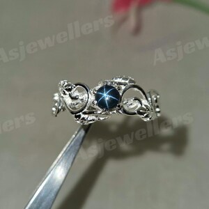 May include: A silver ring with an elaborate design, featuring a dark blue star sapphire gemstone at the centre. The ring is held by tweezers, with the text "Asjewellers" visible in the background.