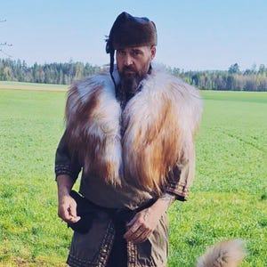 Könnte beinhalten: Ein Mann in einer braunen, pelzbesetzten Jacke und einem schwarzen Hut steht auf einer grünen Wiese mit einem großen braunen und weißen Hund. Der Hund schaut den Mann an und hat die Zunge herausgestreckt.