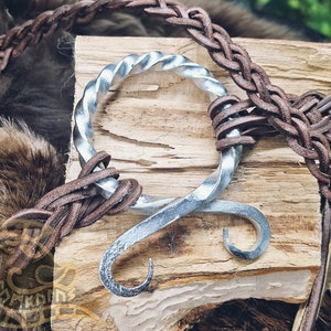 Peut inclure: Un anneau en métal argenté avec un design torsadé, reposant sur un morceau de bois. L'anneau est entouré d'une lanière en cuir marron.