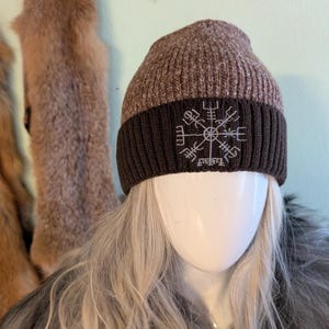 Può includere: Un berretto lavorato a maglia marrone e marrone scuro con un simbolo ricamato bianco. Il cappello ha una trama a coste e un bordo ripiegato. Il berretto è esposto su una testa di manichino con capelli biondi.