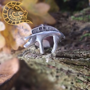 Könnte beinhalten: Ein silberner Schweinchen-Anhänger mit detailliertem Design. Der Anhänger liegt auf einem Stück Holz mit einem verschwommenen Hintergrund aus Herbstblättern.
