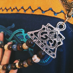 Pode incluir: Um pingente de metal prateado com um design intrincado pendura de um colar de contas. O pingente tem a forma de um triângulo com cantos arredondados e apresenta uma forma de coração estilizada no centro. O colar é feito de contas marrons e brancas.