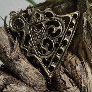 Könnte beinhalten: Ein goldfarbener Metallanhänger mit einem komplizierten Design. Der Anhänger ist dreieckig und hat oben ein Loch für eine Kette.