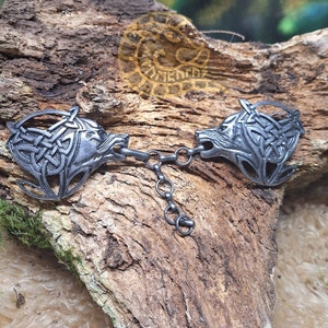 May include: A pair of silver-toned wolf head brooches connected by a chain. Each brooch features a wolf's head with intricate Celtic knotwork designs.