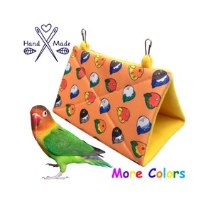 Puede incluir: Una pequeña casita para pájaros naranja y amarilla con un patrón de pájaros coloridos. La casita está hecha de tela y tiene dos lazos para colgar. Un agapornis verde y naranja está sentado frente a la casita.