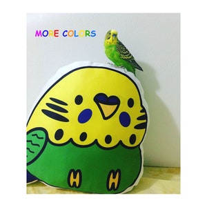 Puede incluir: Una almohada de felpa amarilla y verde con forma de periquito con ojos negros y un pico negro. Un periquito verde está posado encima de la almohada. El texto "MORE COLORS" está escrito en letras de arcoíris sobre la almohada.