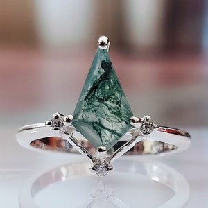 Op de afbeelding: Een zilveren ring met een groene mosagaat edelsteen in een geometrische vorm. De ring heeft kleine heldere stenen aan de zijkanten.